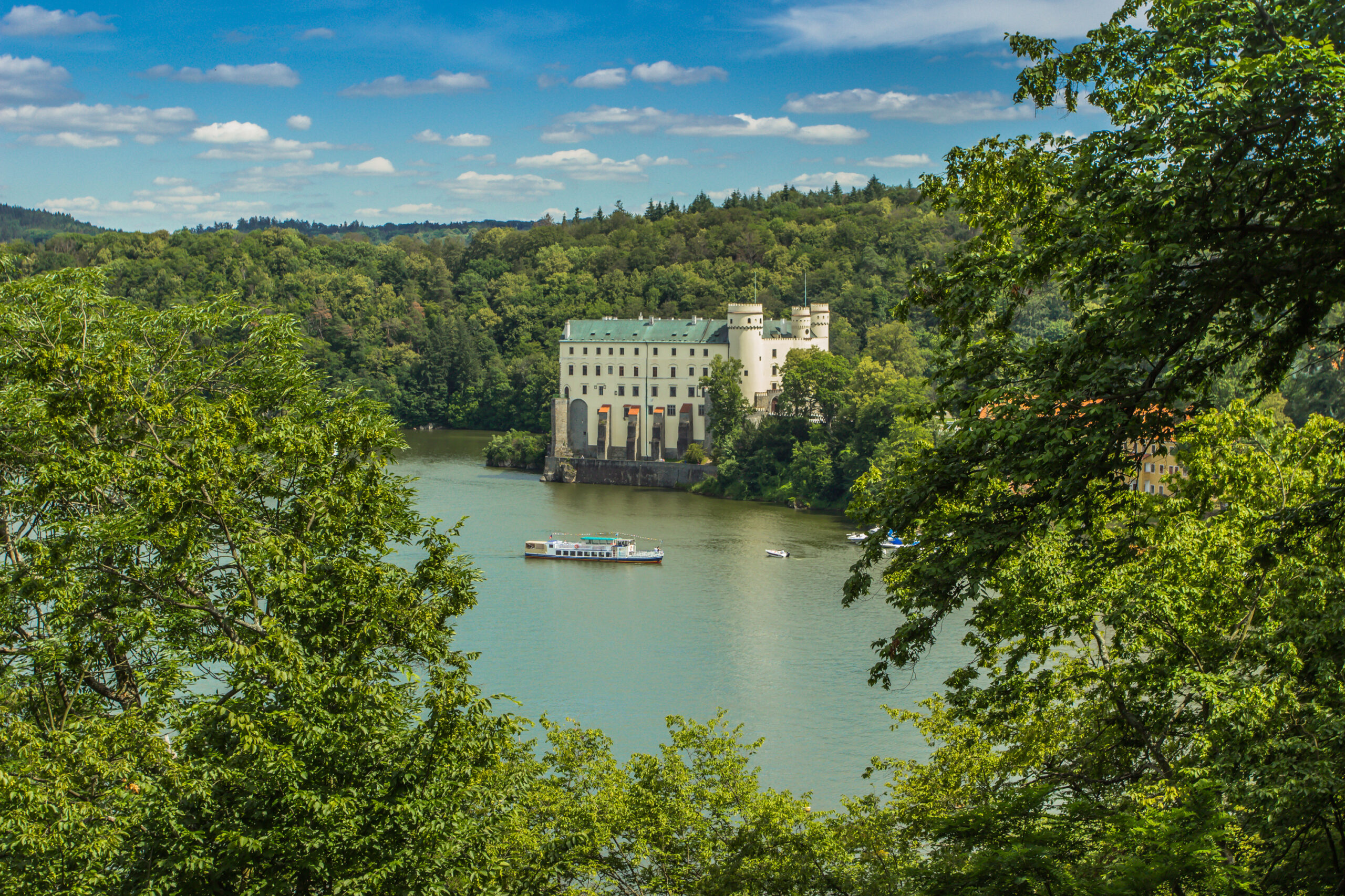 Bílý hrad se zelenými střechami stojí na břehu jezera, obklopený hustým zeleným lesem. Loď pluje po vodě před hradem pod modrou oblohou s roztroušenými mraky.